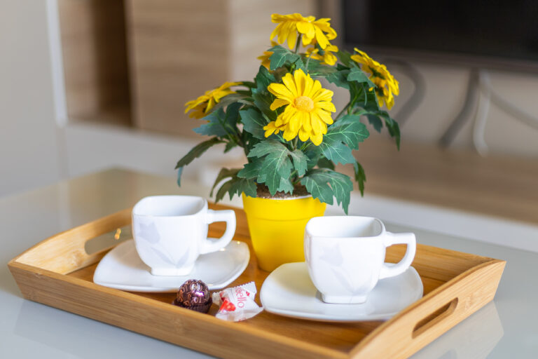 Tazas blancas sobre bandeja de madera, junto a una planta de flores amarillas en maceta amarilla, sobre mesa clara.