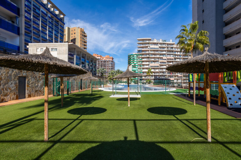 Área de césped artificial con sombrillas de paja, piscina al fondo, edificios modernos y un parque infantil colorido.