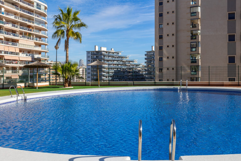 Piscina circular, agua azul brillante, rodeada de edificios altos y una palmera. Cielo despejado, sombrillas de paja.