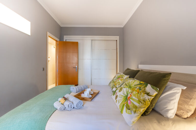 Habitación con cama, cojines de hojas verdes, manta turquesa. Toallas enrolladas y bandeja con tazas sobre la cama. Puerta de madera y armario.