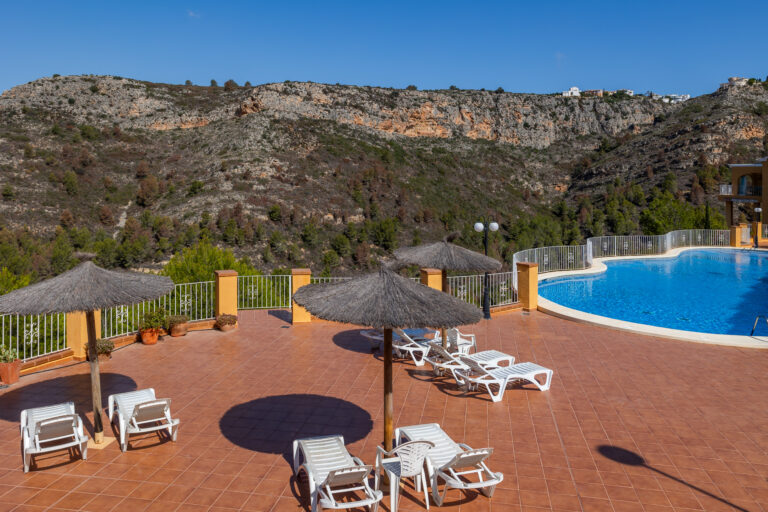 Piscina azul rodeada de tumbonas y sombrillas de paja sobre suelo de azulejos marrones, montañas al fondo.