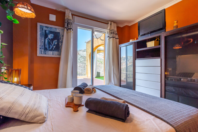 Habitación con paredes naranjas, cama con toallas y tazas, cuadro en la pared, ventana a terraza con vista a montañas.