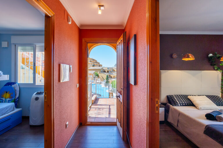 Pasillo con paredes rojas, puertas de habitaciones a los lados, puerta al fondo con vista a piscina y edificios.