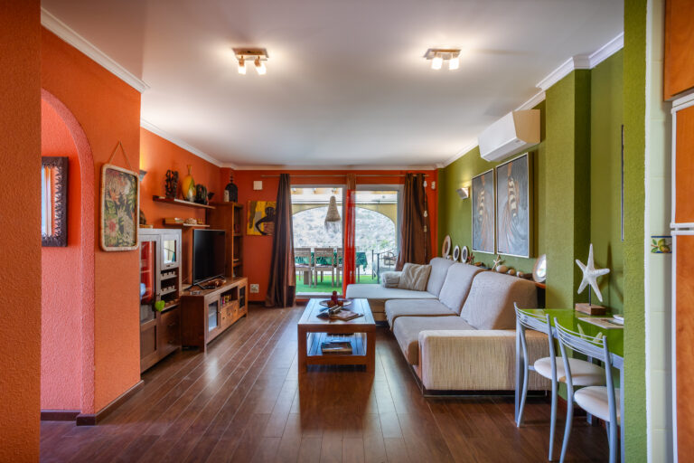 Sala de estar colorida con paredes naranjas y verdes, sofá beige, mesa central, televisión y decoración exótica. Ventana grande al fondo.