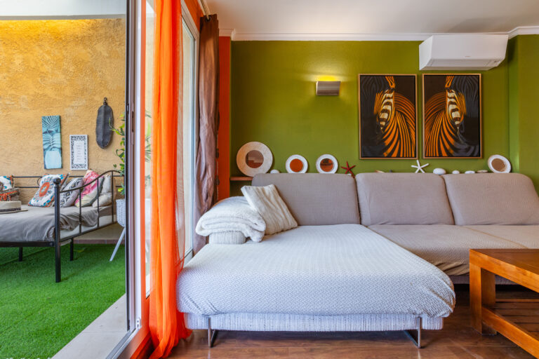 Sala de estar con sofá beige, pared verde con arte abstracto, cortinas naranjas, y terraza con césped artificial.