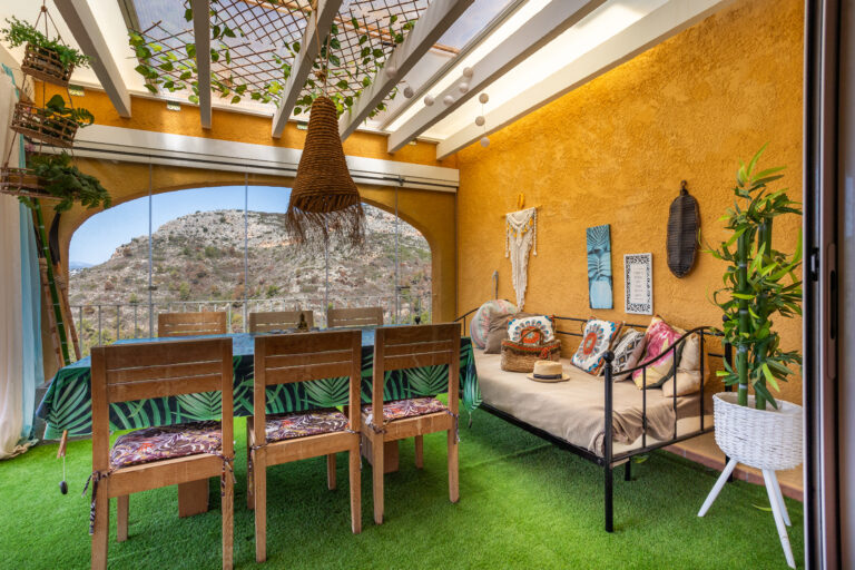 Espacio interior con paredes amarillas, césped sintético, mesa y sillas de madera. Vista panorámica de montañas. Decoración bohemia.