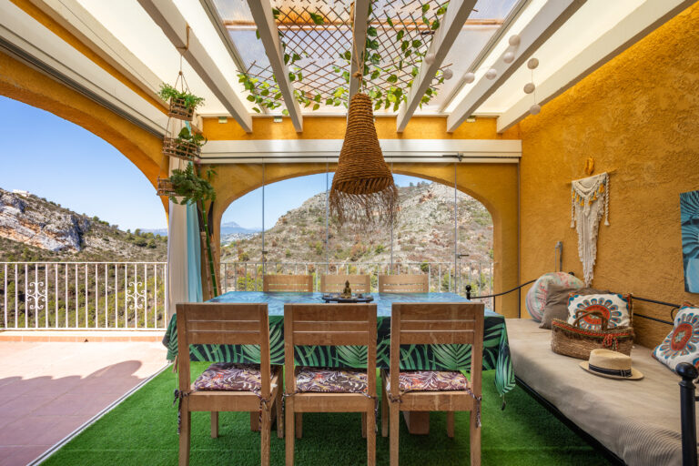 Interior de terraza acristalada, con muebles de madera, mesa decorada, colgante de paja y vistas montañosas.