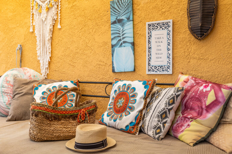 Almohadones con diseños coloridos sobre un sofá beige, sombrero y bolso de paja, pared amarilla con decoraciones.