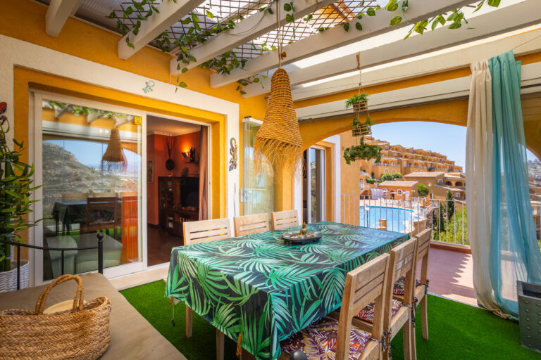 Terraza cubierta con mesa, sillas de madera, mantel de hojas verdes, lámpara colgante, vista a piscina y edificios.