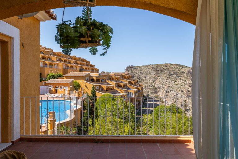 Edificio mediterráneo color ocre con balcones, piscina azul, vegetación verde y montaña rocosa al fondo, bajo cielo despejado.