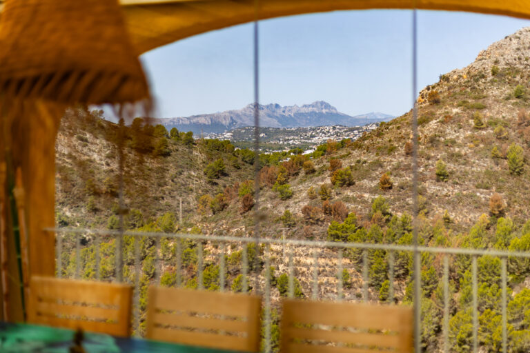 Vista de un valle montañoso con vegetación verde y marrón, sillas borrosas en primer plano, montañas en el horizonte.