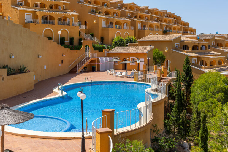 Complejo con edificios amarillos, piscina azul curva y tumbonas. Vegetación y escaleras rodean el área.