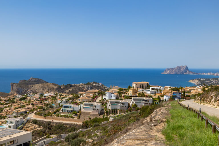 Vista al mar con viviendas blancas y tejados rojos, montañas en el horizonte y vegetación en primer plano.