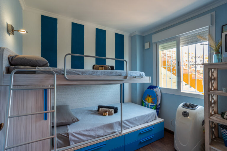 Dormitorio con litera azul, camas grises, pared con franjas azules y blancas. Ventana con luz natural y aire acondicionado portátil.
