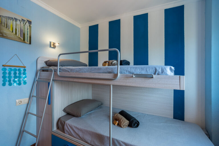 Habitación con literas, pared azul con rayas verticales, cuadro de cabañas, luces, y decoraciones circulares azules.