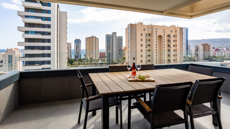 Terraza moderna con mesa de madera, copas de vino, y aperitivos. Skyline urbano y mar en el fondo bajo cielo parcialmente nublado.