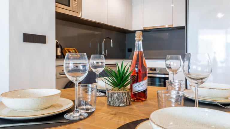 Botella de vino rosado, copas de vino, y platos en una mesa de madera. Cocina moderna con electrodomésticos en el fondo.