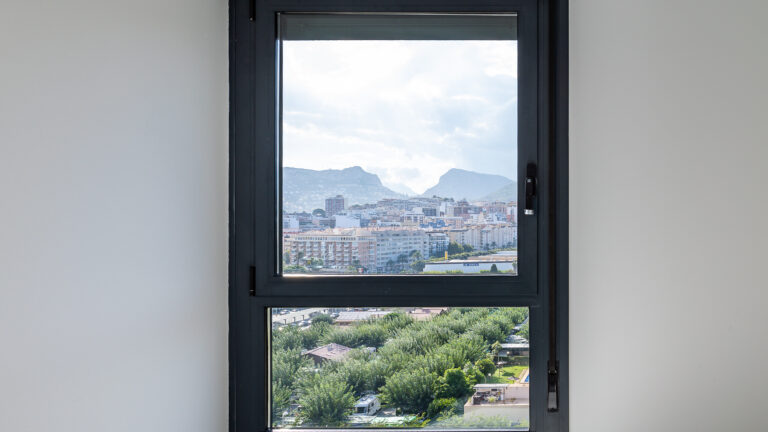 Vista desde ventana: ciudad con edificios, montañas al fondo y vegetación en primer plano bajo cielo parcialmente nublado.