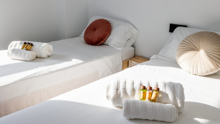 Habitación con dos camas individuales, cojines redondos en tonos tierra, toallas blancas y amenidades sobre las camas.