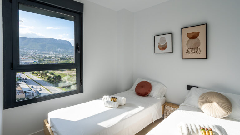 Habitación luminosa con dos camas individuales, cojines redondos, cuadros abstractos, ventana con vista a montañas y ciudad.