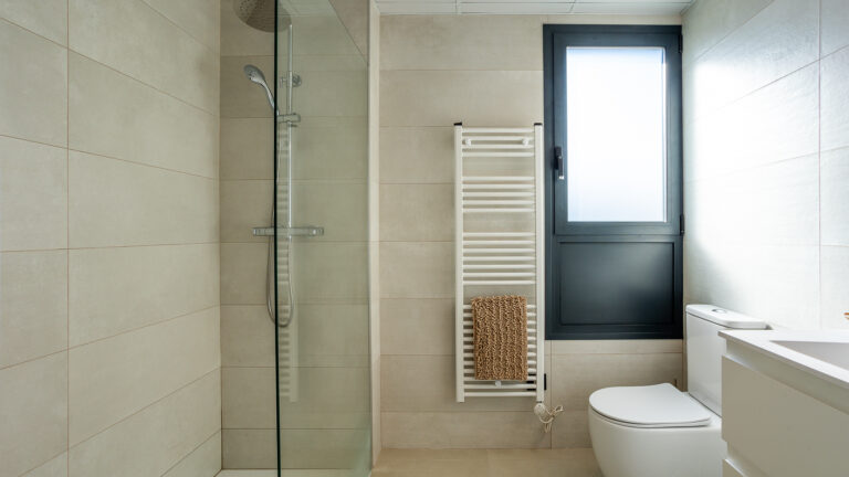 Baño moderno con ducha de cristal, inodoro blanco, calentador de toallas y ventana con marco oscuro. Baldosas beige.