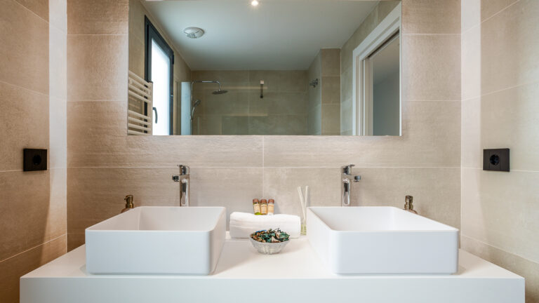 Baño moderno con dos lavabos blancos, grifos metálicos, espejo grande, toallas blancas y artículos de tocador. Paredes beige.