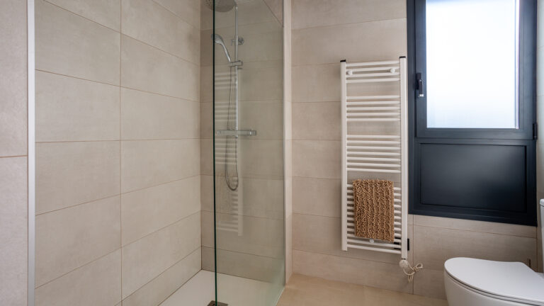 Baño moderno con ducha de vidrio, azulejos beige, toalla en radiador blanco, ventana oscura y váter blanco.