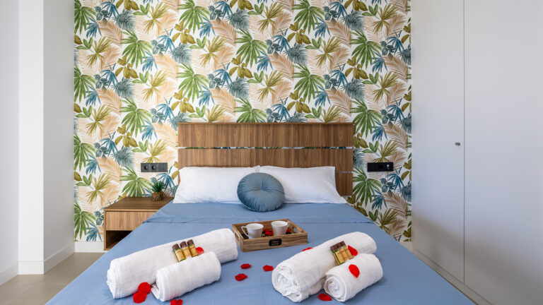 Cama con sábanas azules, cabecero de madera, fondo de papel tapiz tropical, toallas blancas y pétalos rojos.