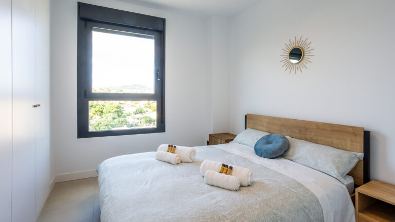 Dormitorio con cama doble, ropa de cama azul claro, cojines, ventana amplia con vista y espejo decorativo en la pared.