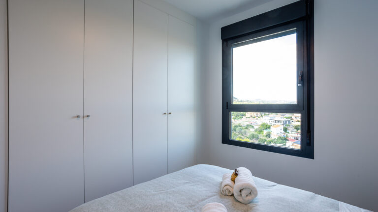 Habitación con armario blanco, ventana grande con vista panorámica, cama con sábanas grises y toallas enrolladas sobre ella.