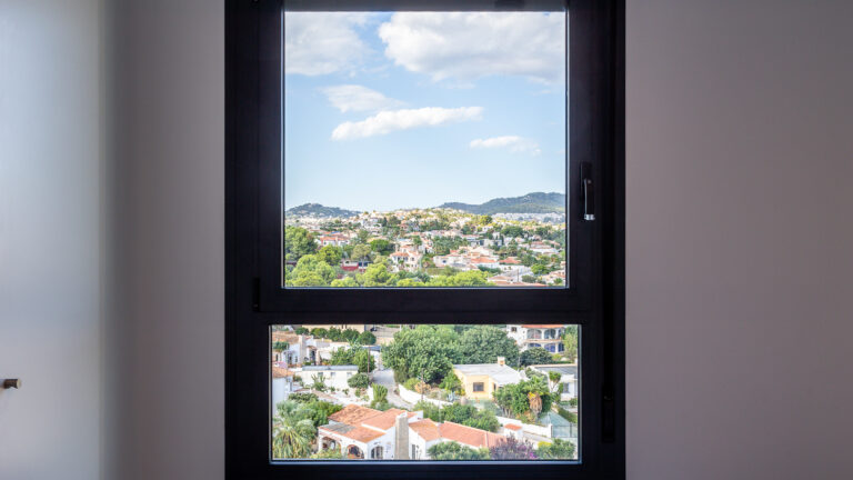 Vista desde una ventana mostrando un paisaje urbano con casas entre árboles, colinas y cielo azul con nubes.