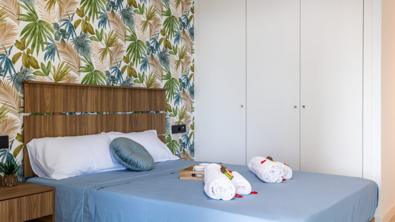 Habitación con cama de sábanas azules, cabecero de madera, y pared con papel tapiz de hojas tropicales en tonos verdes y marrones.