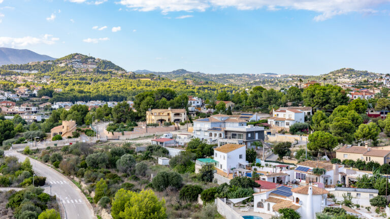 Vista panorámica de casas en una colina arbolada, con montañas al fondo y un cielo azul claro.