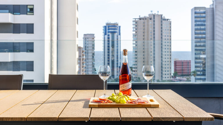Mesa con botella de vino rosado, copas, uvas y embutidos. Fondo de edificios altos y vista al mar.