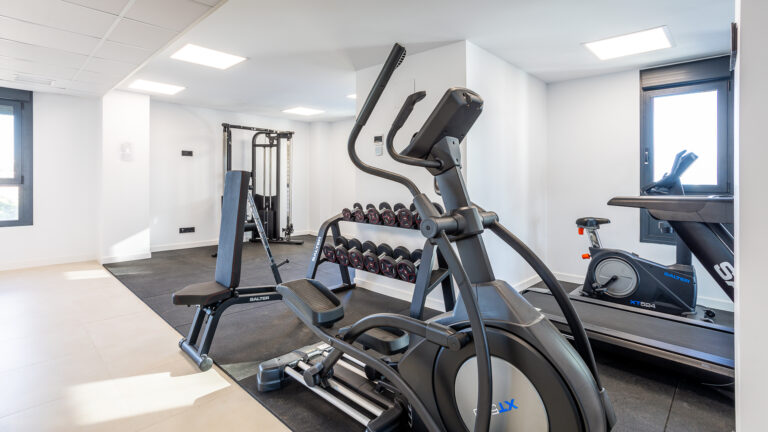 Gimnasio moderno con elíptica, pesas, banco de ejercicios, bicicleta estática y cinta de correr en un ambiente luminoso.