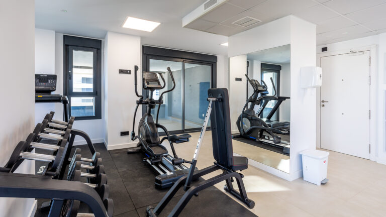 Gimnasio pequeño con máquinas, pesas en estantería, espejo grande, ventana y puerta blanca. Espacio limpio e iluminado.