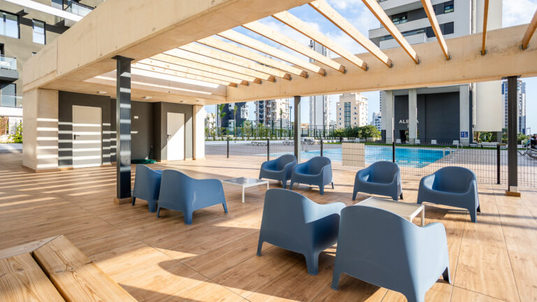 Terraza con techo de vigas de madera, sillas azules, mesas, junto a una piscina, rodeada de edificios modernos.
