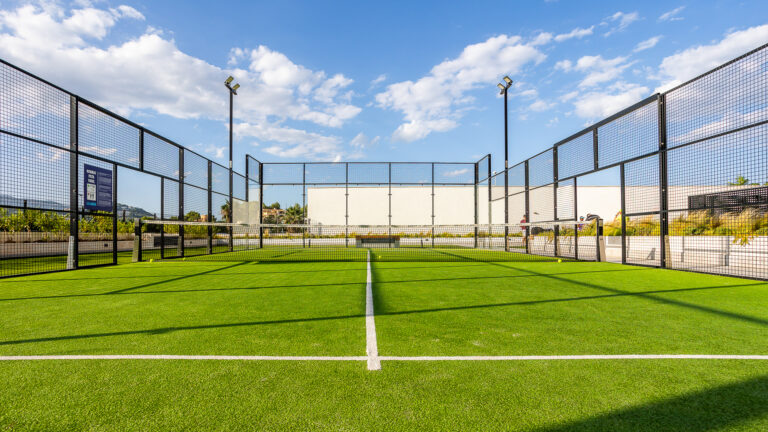 Cancha de pádel verde con rejas de metal, red central y fondo con cielo azul y nubes blancas.