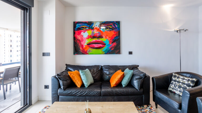 Sala moderna con sofá de cuero negro, cojines naranjas y verdes. Pintura vibrante de un rostro en la pared blanca. Ventana a la izquierda.