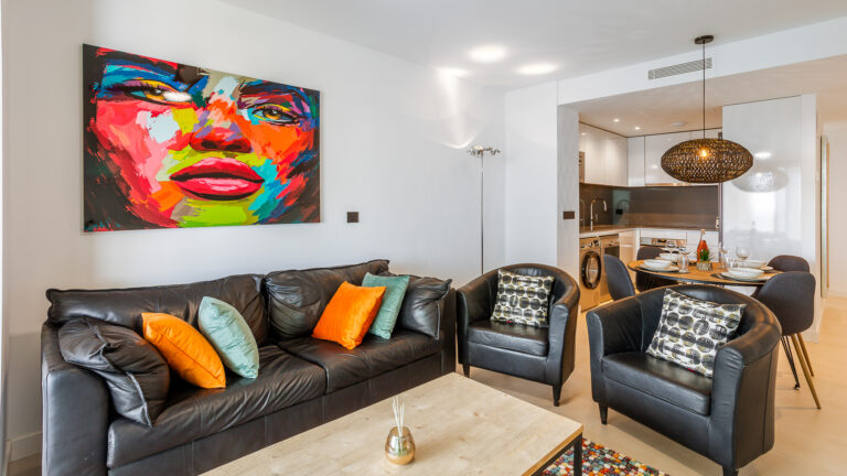 Sala moderna con sofá de cuero negro, almohadas naranjas y verdes. Pintura colorida en la pared, junto a cocina abierta y comedor.