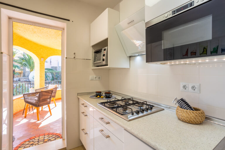 Cocina moderna con encimera beige, estufa de gas, microondas y salida a terraza con sillas de mimbre bajo un arco amarillo.