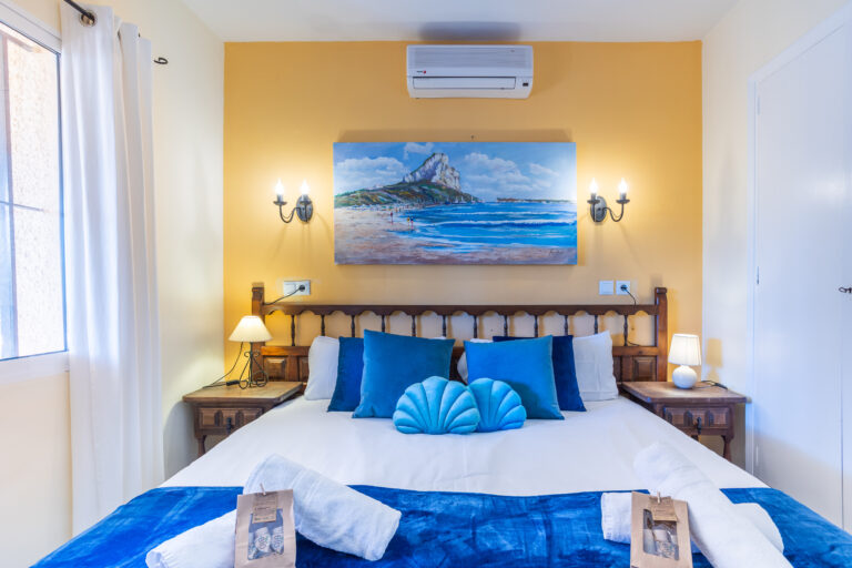 Habitación con cama doble, almohadas azules, cuadro de playa, lámparas de mesa, pared amarilla y aire acondicionado.