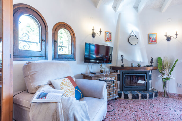 Sala acogedora con chimenea, sillón beige, ventanas arqueadas, televisor en la pared, y decoración con cuadros y plantas.