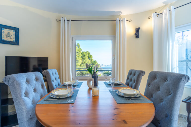 Comedor con mesa de madera, cuatro sillas grises, ventana al fondo con vista a árboles, platos y copas sobre manteles individuales.