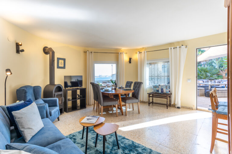 Sala de estar luminosa con sillones azules, mesa de comedor redonda, televisor y estufa. Ventanas con cortinas blancas, vista al exterior.