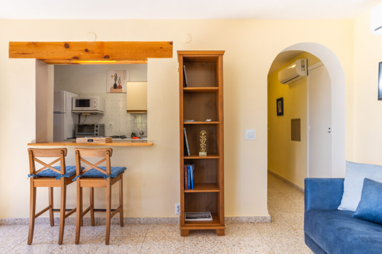 Sala con barra de madera, dos sillas con cojines azules, estantería, cocina al fondo y sofá azul a la derecha.