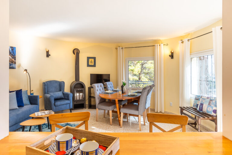 Sala de estar con sillones azules, mesa de comedor redonda, estufa, ventanas amplias y cortinas blancas. Ambiente cálido y acogedor.