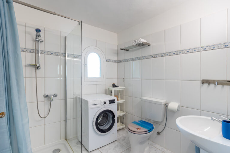 Baño con azulejos blancos, ducha con cortina azul, lavadora, inodoro con tapa azul, y pequeño fregadero blanco.