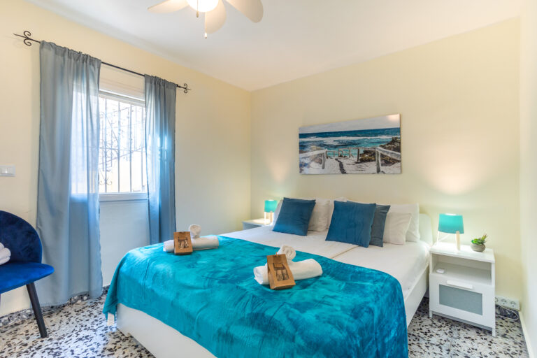 Dormitorio luminoso, cama doble con ropa de cama azul, lámparas de mesa, cuadro de playa y cortinas azules. Suelo de mosaico.
