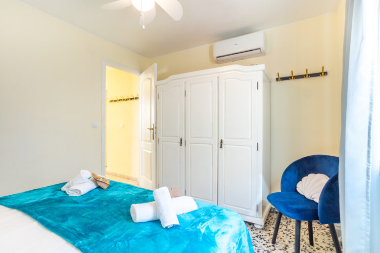 Habitación con cama, colcha azul, toallas blancas, armario blanco, aire acondicionado, silla azul y perchero en la pared.